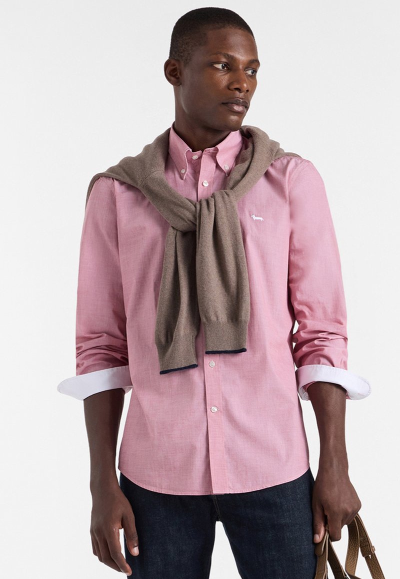 Camicia button-up rosa chiaro con maniche arrotolate, abbinata a un maglione marrone drappato intorno al collo. Jeans scuri e una borsa marrone completano l'outfit.
