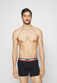 Melni bokseri ar mīkstu elastīgu jostu, kas rotāta ar sarkaniem un baltajiem svītrainiem un "Levi's" uzrakstu. Modelis stāv uz neitrāla fona.