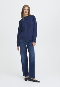 Sieviete, kura ģērbusies tumši zilā džemperī, plaša piegriezuma tumšzilās biksēs un melnās zemas papēža kurpēs, stāvot uz vienkārša balta fona.