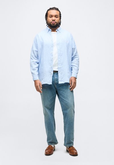 Άνδρας που στέκεται με το πρόσωπο προς τα εμπρός, φοράει γαλάζιο ριγέ πουκάμισο με κουμπιά, λευκό t-shirt, μπλε τζιν και καφέ δερμάτινα μοκασίνια.