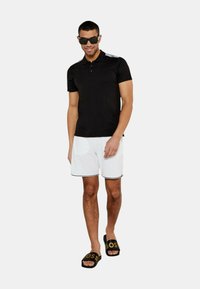 Polo nero con polo a tre bottoni, dettagli a righe bianche sulle spalle, abbinato a pantaloni corti bianchi e sandali neri con logo dorato.