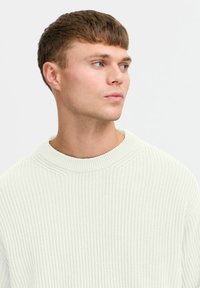 Hvit ribbestrikket genser med en bølget tekstur og vid, rund halslinning. Stoffet ser mykt og litt elastisk ut.