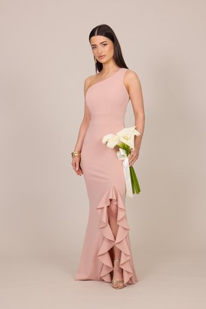 Frau in einem einseitigen, blassrosa Kleid mit Rüschendetail, die weiße Calla-Lilien hält, welche mit einem weißen Band gebunden sind, stehend vor einem neutralen Hintergrund.