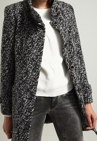 Uldblandingsfrakke i sort og hvid tweedmønster, med høj krave og brune knapper. Matcher med en hvid strikket sweater og grå jeans.