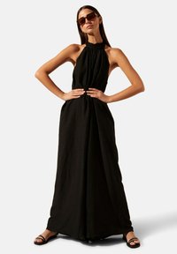 Robe maxi noire à bretelles, avec un col froncé, un tissu fluide et une coupe décontractée. Portée avec des lunettes de soleil foncées et des sandales plates.