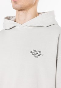 Felpa grigia chiara in cotone con una vestibilità rilassata, dotata di una tasca a marsupio, cappuccio con coulisse e logo ricamato sul petto.