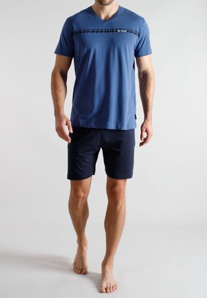 Mann trägt blaues V-Ausschnitt-T-Shirt und dunkle Shorts, steht barfuß vor einem einfachen hellen Hintergrund.