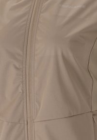 Beige atletisk jacka med en slät yta, framdragkedja och diskret logotyp på bröstet för en funktionell design.