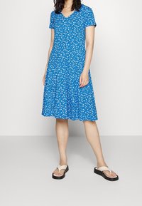 Femme portant une robe bleue à fleurs arrivée au genou et des tongs en plateforme beige se tenant devant un fond blanc uni.
