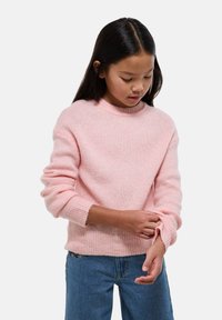 Fille aux longs cheveux noirs ajustant la manche d'un pull en tricot rose clair, portant un jean bleu, sur un fond clair.