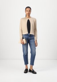 Beige neulepusero, jossa on pyöreä kaulus, lyhyt malli ja logo-detailit helman alueella. Yhdistetty vaaleansinisiin suoriin farkkuihin ja mustiin loafer-kenkiin.