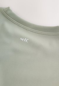 Világoszöld sportos felső lekerekített nyakkivágással, sima textúrával, elöl fehérrel nyomtatott "self." márkanévvel.