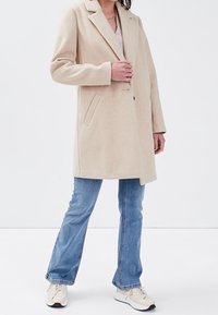 Beige uldblandingsfrakke med slagkrave, enkeltknaplukning, to sidelommer og lige snit, parret med blå jeans og sneakers.