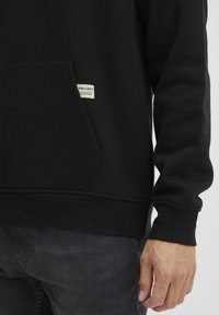 Sudadera negra con un bolsillo frontal, puños y dobladillo acanalados. Presenta una pequeña etiqueta blanca con "PROJECT" y "PUBLISHED 2020."