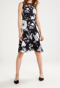 Svart klänning med vita blommönster, ärmlös design och genomskinliga detaljer på överkroppen; nederkanten slutar strax ovanför knäna. Svarta skor bärs.