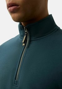 Ciemnozielony polar na zamek z wysokim kołnierzem, z metalowym zamkiem błyskawicznym i metką marki. Materiał wydaje się gładki z delikatnym połyskiem.