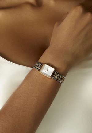 Montre rectangulaire argentée et dorée portée au poignet, présentant un cadran blanc, de fins marqueurs d'heure, et un bracelet en métal tissé avec des maillons d'accent.
