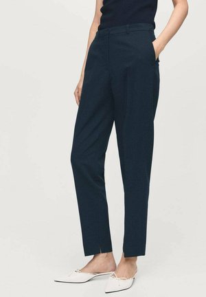 Donna che indossa pantaloni tapered blu navy con pieghe frontali e ballerine bianche con punta, in piedi con una mano in tasca contro uno sfondo semplice.