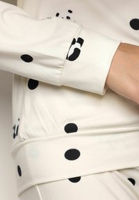 Tessuto color crema con pois neri, caratterizzato da maniche lunghe con polsini e una vita aderente. Texture morbida e elementi di design minimal.