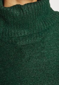 Maglione a collo alto in maglia verde con texture a coste. Il tessuto appare morbido e testurizzato, mostrando una tonalità verde profondo.