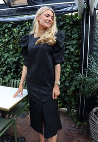 Rochie midi neagră cu buline, mâneci bufante, despicătură frontală și țesătură netedă. Purta cu un ceas de mână. Frunziș verde în fundal.