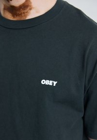 Black cotton t-shirt featuring a round neck, short sleeves, and a white "OBEY" logo on the left side of the chest. Textured fabric.