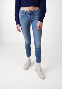 Ljusblå skinny jeans med midja i normalhöjd, urtvättad finish och subtila gnuggningar. Bärs med vita strumpor och bruna sneakers.