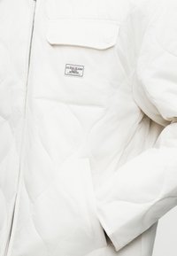 Baltas siuvinėtas švarkelis su užtrauktuku ir krūtinės kišene bei prekės ženklo etikete. Audinys yra lygus ir tekstūruotas.