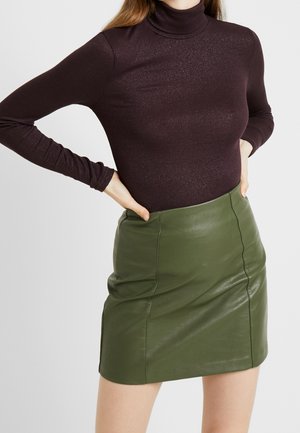 Mujer con un suéter de cuello alto de color púrpura oscuro y una minifalda de cuero verde oliva, con las manos en la cintura.