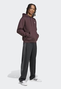 Burgunderfarget hettegenser med frontlomme; kombinert med svarte bukser med hvite striper nedover sidene. Hvite treningssneakers.