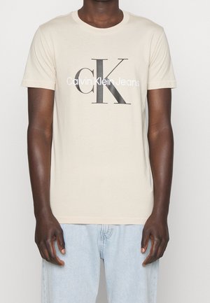 Mand iført en beige t-shirt med stor sort tekst "cK" og hvid tekst "Calvin Klein Jeans", kombineret med lyseblå jeans.