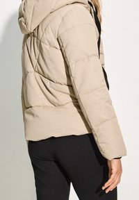 Beige, kviltet pufferjakke med hætte, der har en glat overflade og en lynlåslomme på siden. Sorte detaljer nær lynlåsen.