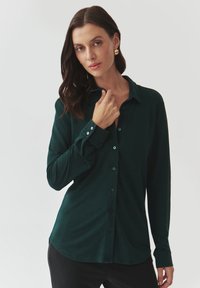 Šedozelená dlouhá košile s knoflíky a límečkem, vyrobená z hladkého materiálu a s kontrastními knoflíky. Model jemně sahá na límeček.