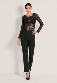 Vrouw staat met hand op de heup, gekleed in een zwarte doorschijnende kanten top met lange mouwen, een zwarte bh, zwarte broek met stippen en zwarte hakken.