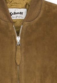 Chaqueta de ante marrón con cremallera plateada parcialmente abierta, mostrando el forro interior acolchado y la etiqueta blanca de Schott NYC en el cuello.