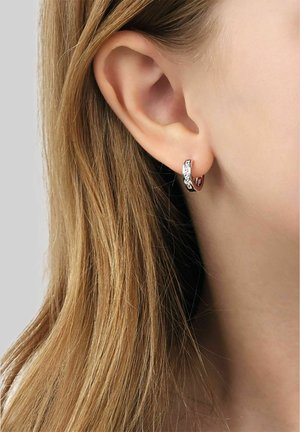 Close-up of a person’s ear with light brown hair, wearing a small silver hoop earring with intricate engraved patterns.