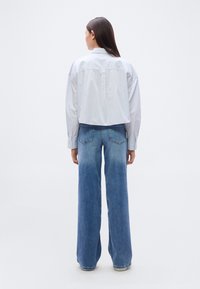 Κοντοκομμένο, ανοιχτό μπλε πουκάμισο με ρίγες και μακριά μανίκια, συνδυασμένο με καμπάνα, ξεθωριασμένο μπλε τζιν παντελόνι. Μινιμαλιστικός σχεδιασμός, χωρίς ορατά λογότυπα.