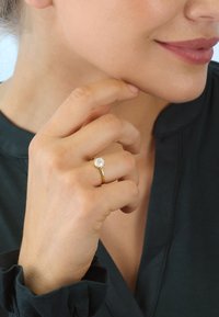 Gold ring with a round, clear stone, worn on a hand with light skin. Dark green blouse with ruffled cuffs, soft lighting.