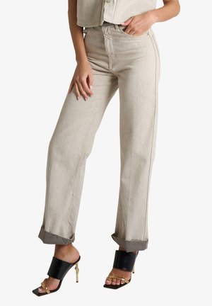 Pantaloni beige a vita alta e gamba larga con risvolti, indossati con sandali aperti neri e dorati con tacco alto.