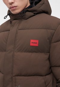 Gros plan d'une personne portant une veste matelassée marron avec capuche et un patch logo rouge "HUGO" sur la poitrine.
