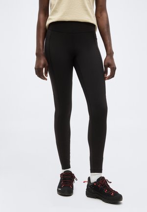 Žena v čiernych priliehavých legínach, čiernej turistickej obuvi s červenými šnúrkami a bielych ponožkách stojaca pred jednofarebným pozadím.