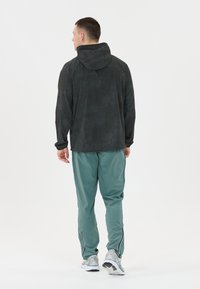 Schwarze Kapuzenjacke mit einem strukturierten Muster, kombiniert mit türkisfarbenen Hose mit reflektierenden Akzenten und Reißverschlüssen, getragen mit hellgrauen Sneakers.