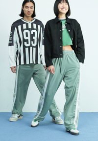 Duas modelos estão em pé sobre um chão azul. Uma está vestida com um casaco preto e uma camisola verde cropped; a outra veste uma camisola às riscas pretas e brancas com calças cinzas.