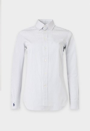 Žena stojaca s rukami v vreckách, oblečená v pruhovanej bielej košeli na gombíky, tmavomodrom tričku, modrých širokých džínsoch a bielych teniskách.