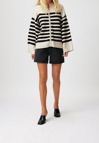 Mujer con un suéter de rayas blancas y negras con cremallera, pantalones cortos de mezclilla negros y mule negros de tacón, de pie sobre un fondo claro liso.