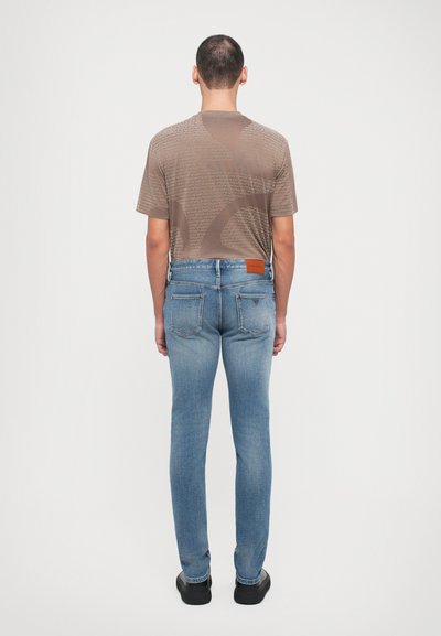 Barna mintás póló és kék, slim-fit farmernadrág bőrdísszel. A farmer textúrája farmeres, míg a póló anyaga sima és könnyű.