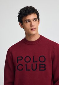Roter Strickpullover mit einem strukturierten Muster, der "POLO CLUB" in großen schwarzen Buchstaben auf der Brust trägt und einen gerippten Halsausschnitt hat.