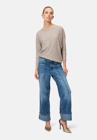 Hellgraues, strukturiertes Oberteil mit dreiviertel Ärmeln; blaue Jeans mit weitem Bein und aufgeschlagenen Säumen; kombiniert mit spitzen goldfarbenen Absätzen.