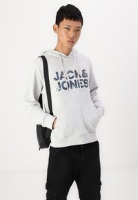 Grauer Hoodie mit "JACK & JONES" Grafik in Blautönen, Kängurutasche, Kapuze mit Zugband, kombiniert mit schwarzen Hose und einem schwarzen Rucksack.
