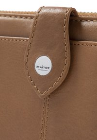 Portefeuille en cuir marron avec une texture lisse, présentant un logo circulaire argenté, des accents cousus et une fermeture à onglet en haut.